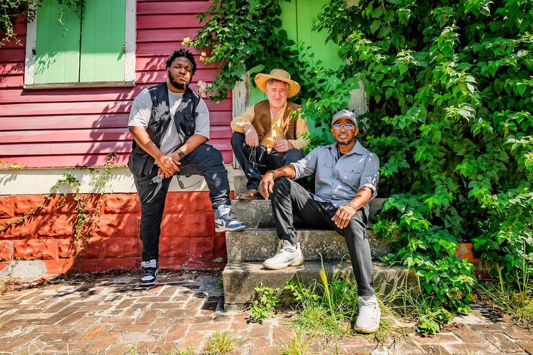 Jon Cleary & The Absolute Monster Gentlemen posing outside a vibrant New Orleans style house surrounded by lush greenery.